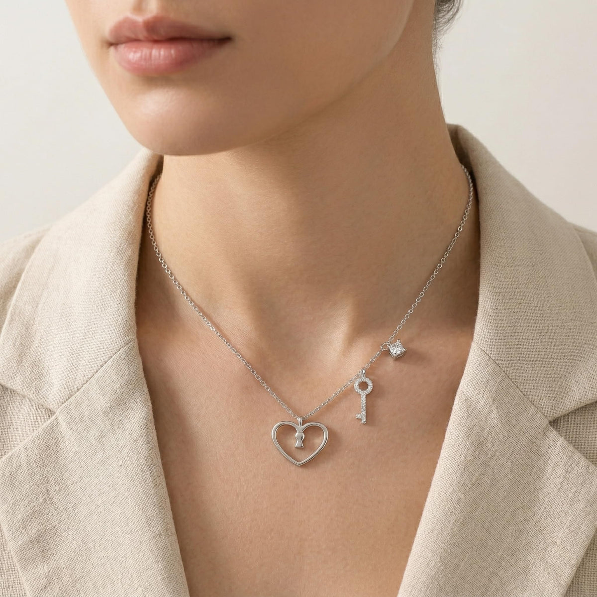 A close-up lifestyle shot of a model wearing a delicate silver necklace with a heart and key pendant, styled with a cream satin top against a warm beige background.