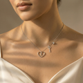 A close-up lifestyle shot of a model wearing a delicate silver necklace with a heart and key pendant, styled with a cream satin top against a warm beige background.
