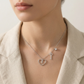 A close-up of a model wearing a delicate silver necklace with a heart-shaped pendant, a key pendant, and a small star charm, styled with a beige linen blazer.