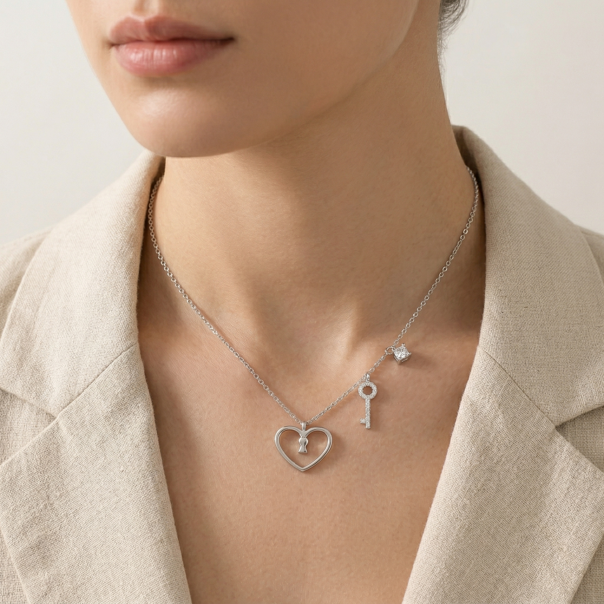 A close-up of a model wearing a delicate silver necklace with a heart-shaped pendant, a key pendant, and a small star charm, styled with a beige linen blazer.