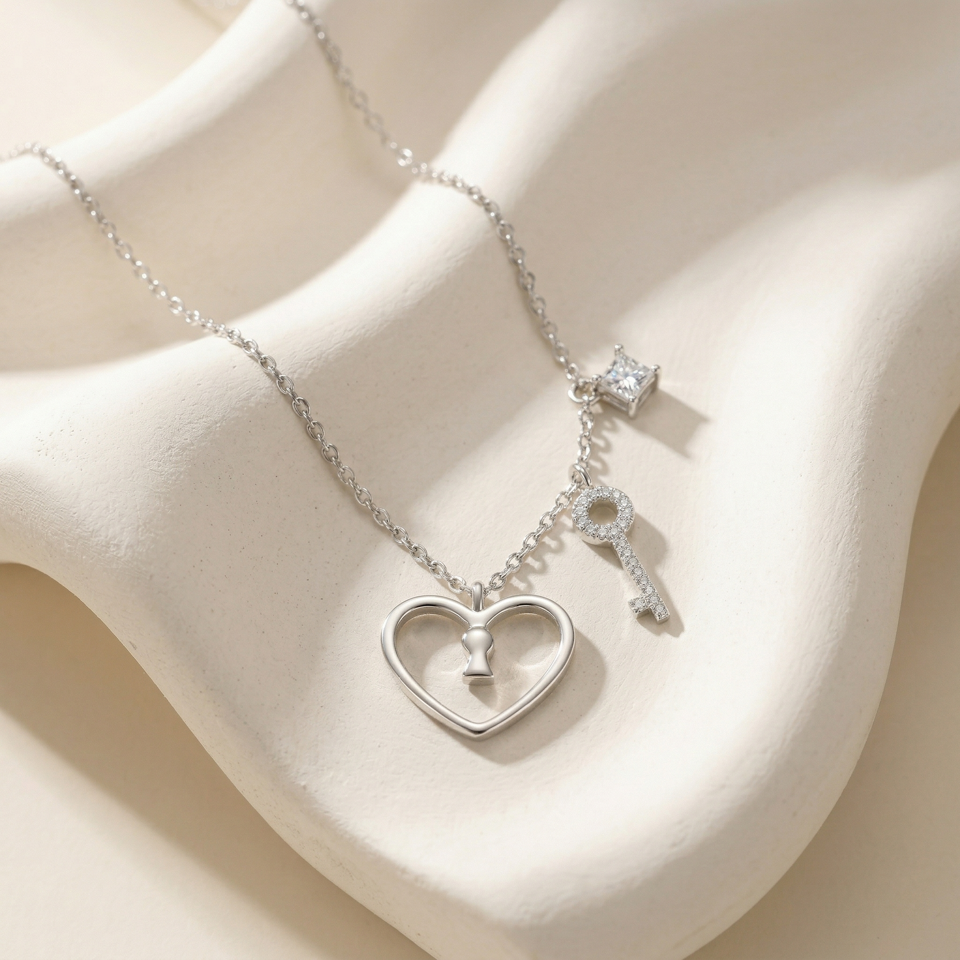 A silver necklace with a heart-shaped pendant containing a small key, a keychain, and a crown charm, displayed on a cream-colored wavy jewelry stand.